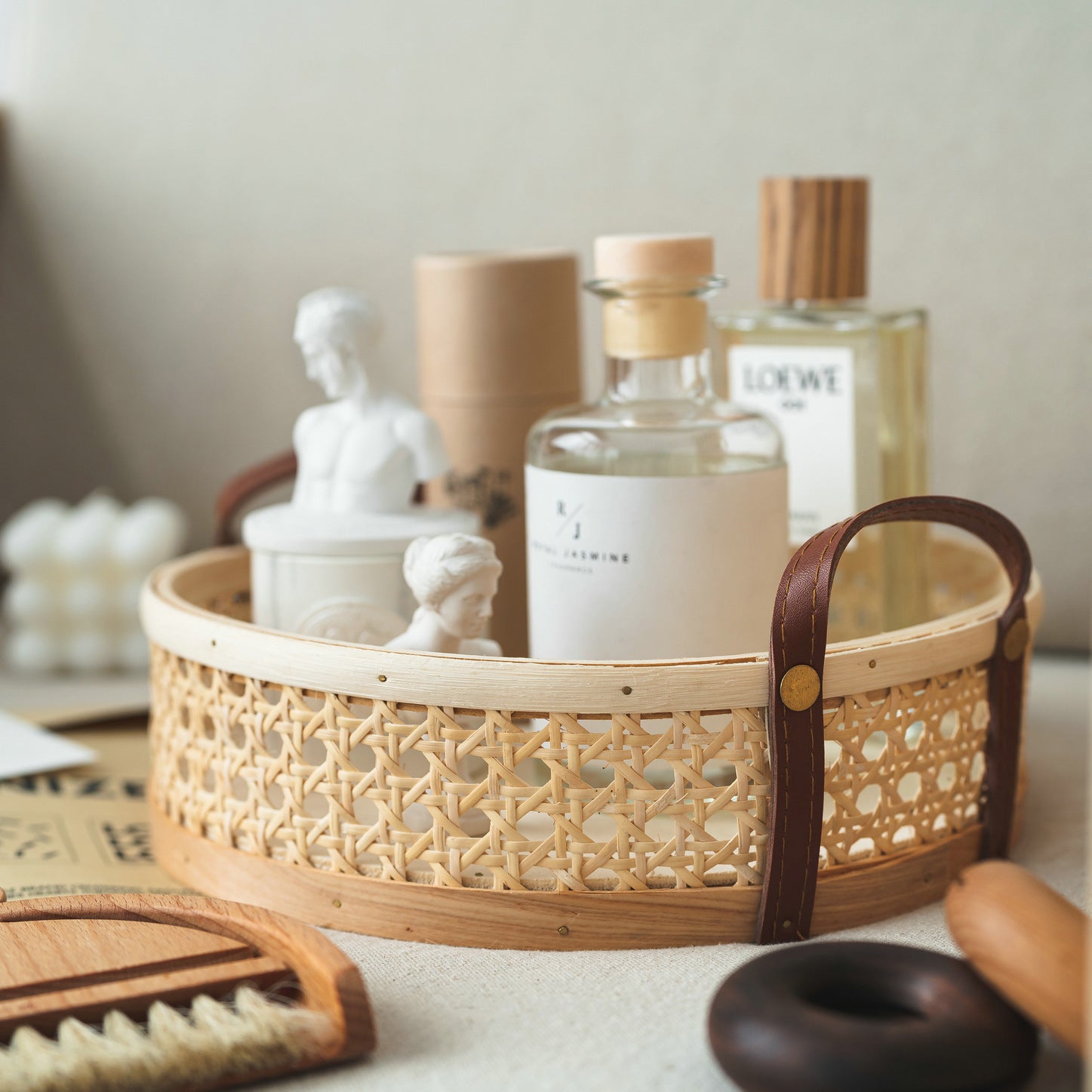 Round wicker tray in natural wood color, placed on an entryway table, organizing keys, perfume, and small items, with convenient leather handles and multiple size options.