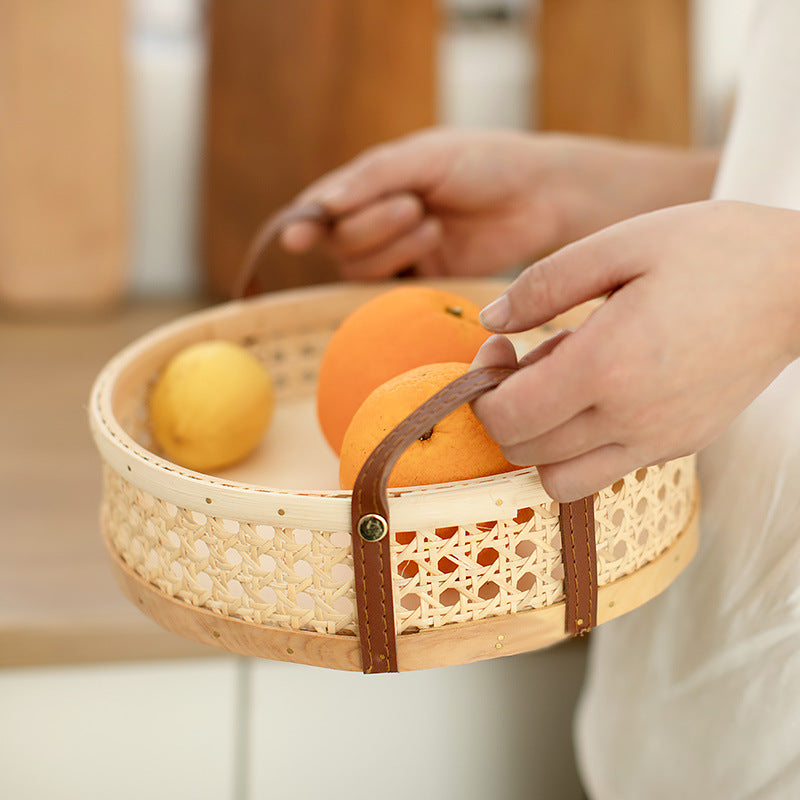 Round rattan wicker storage tray in natural wood color, filled with fresh fruits, showcasing its sturdy leather handles and multiple size options.