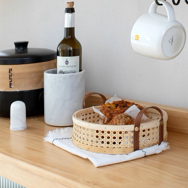 Round rattan wicker storage tray with leather handles, holding fresh bread on a kitchen countertop, available in natural and walnut wood colors, showcasing its durable and eco-friendly design in multiple sizes.