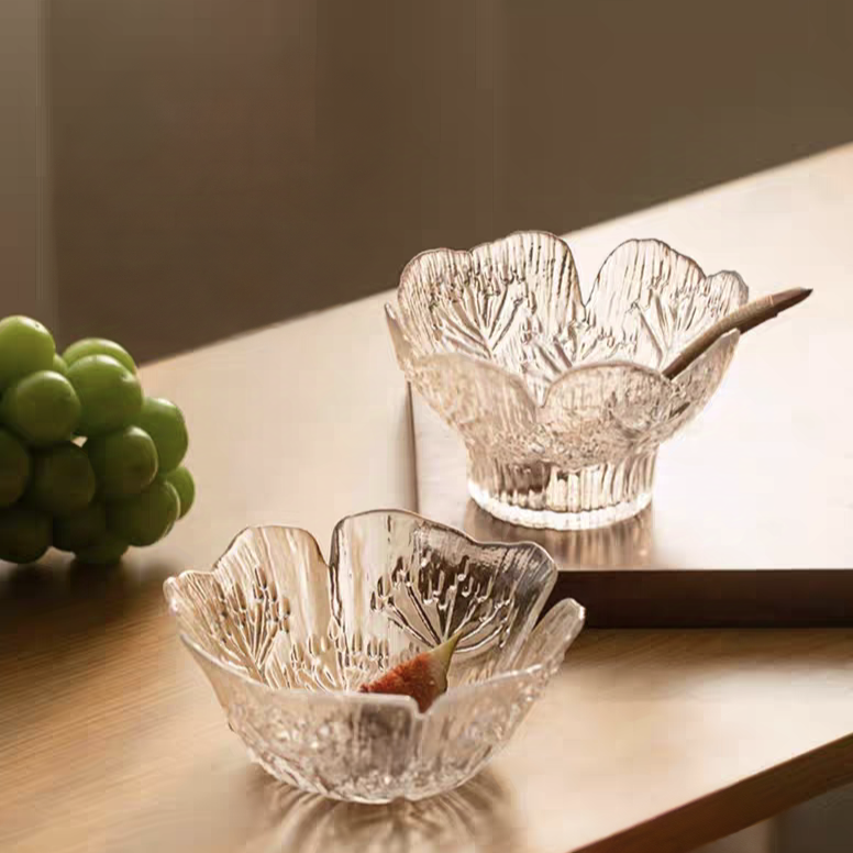 A clear glass dessert bowl with a sleek flat base and petal design, and a high-footed glass bowl with intricate floral embossing, perfect for salads and yogurt in Scandinavian-inspired homes.